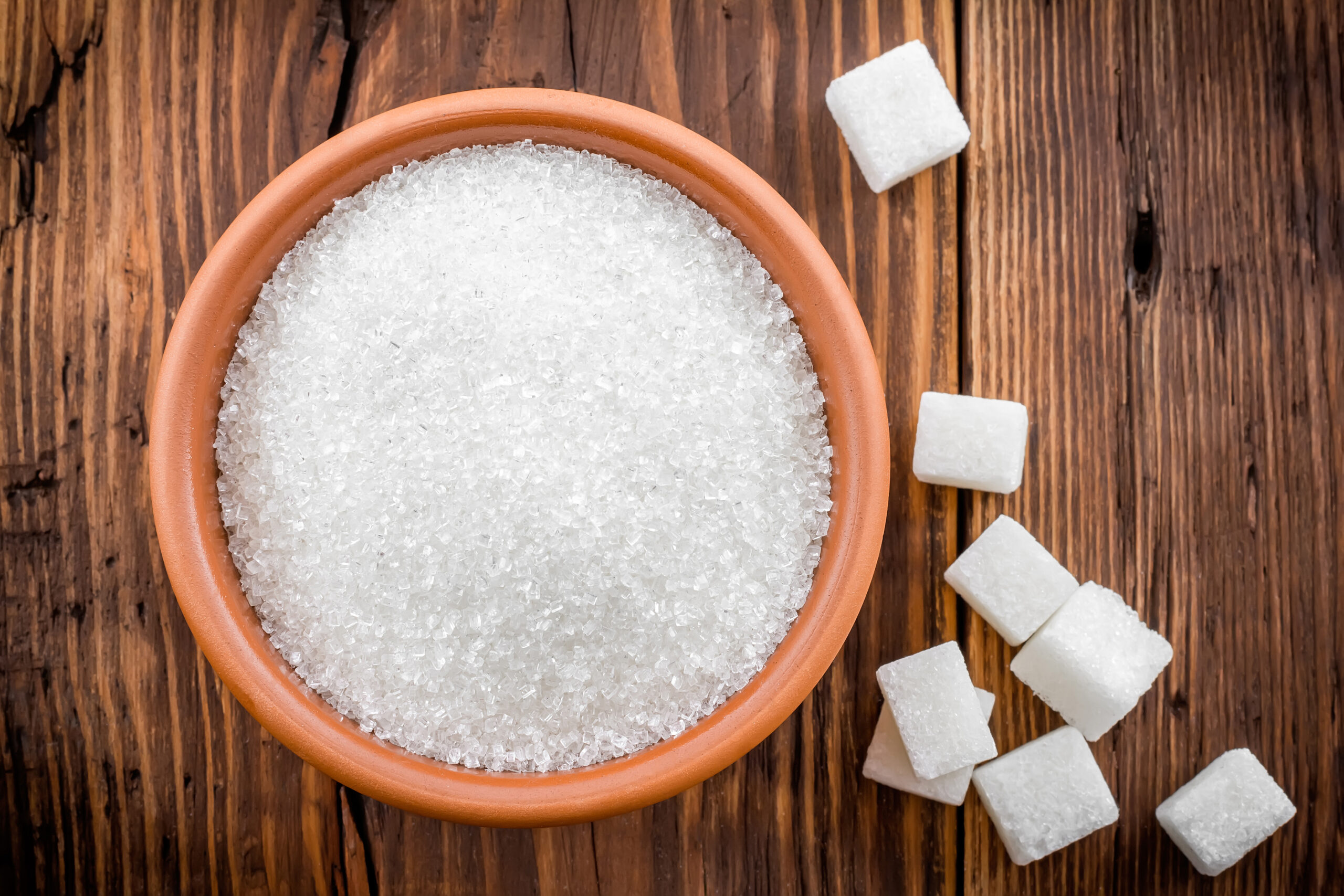 A terracotta bowl filled with coarse white sugar granules on a rustic wooden table, surrounded by scattered sugar cubes.